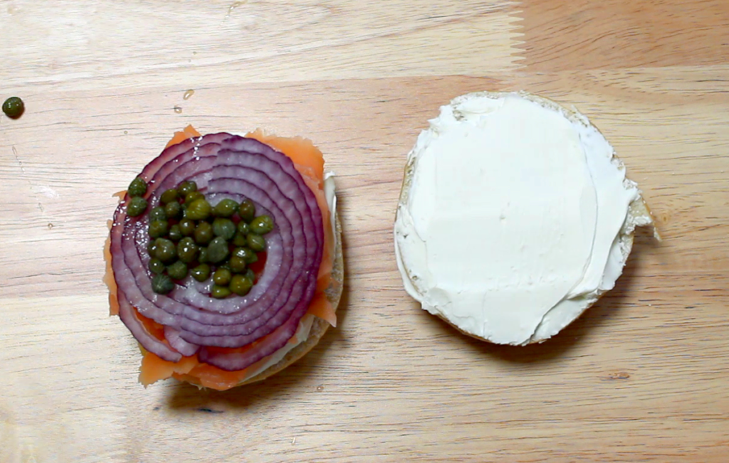 ingredients layered on smoked salmon bagel, cream cheese on one half and cream cheese, salmon, red onion and capers on other half of bagel