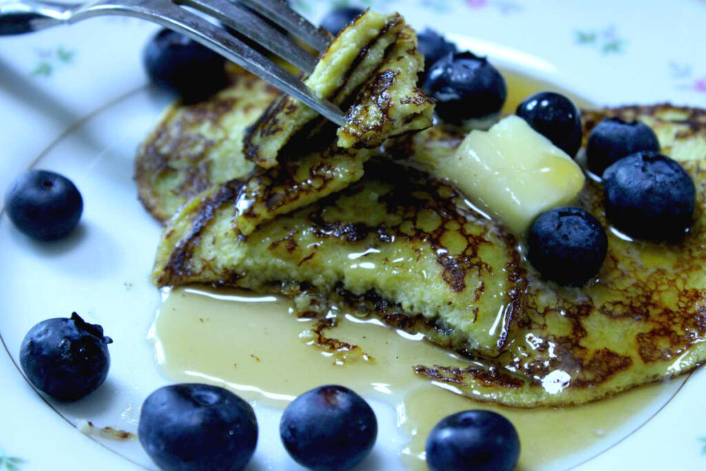 banana egg pancakes on a plate with blueberries and butter covered with maple syrup and a slice cut out on a fork