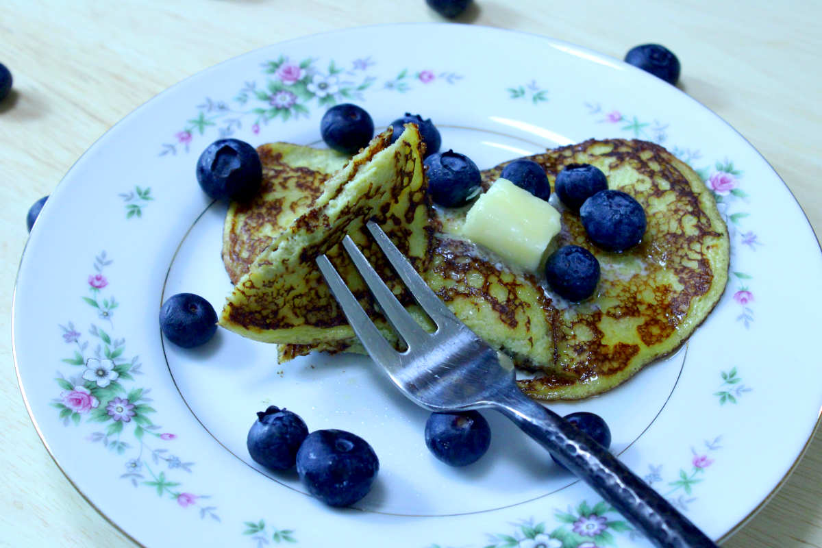 banana egg pancakes on a plate covered with blueberries and butter with a piece cut out on a fork