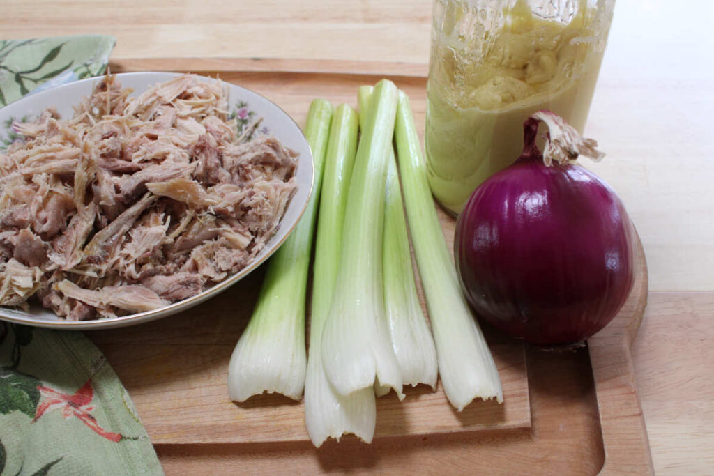 a cutting board with a bowl of shredded chicken or rabbit with celery stalks, a red onion and a jar of homemade mayo