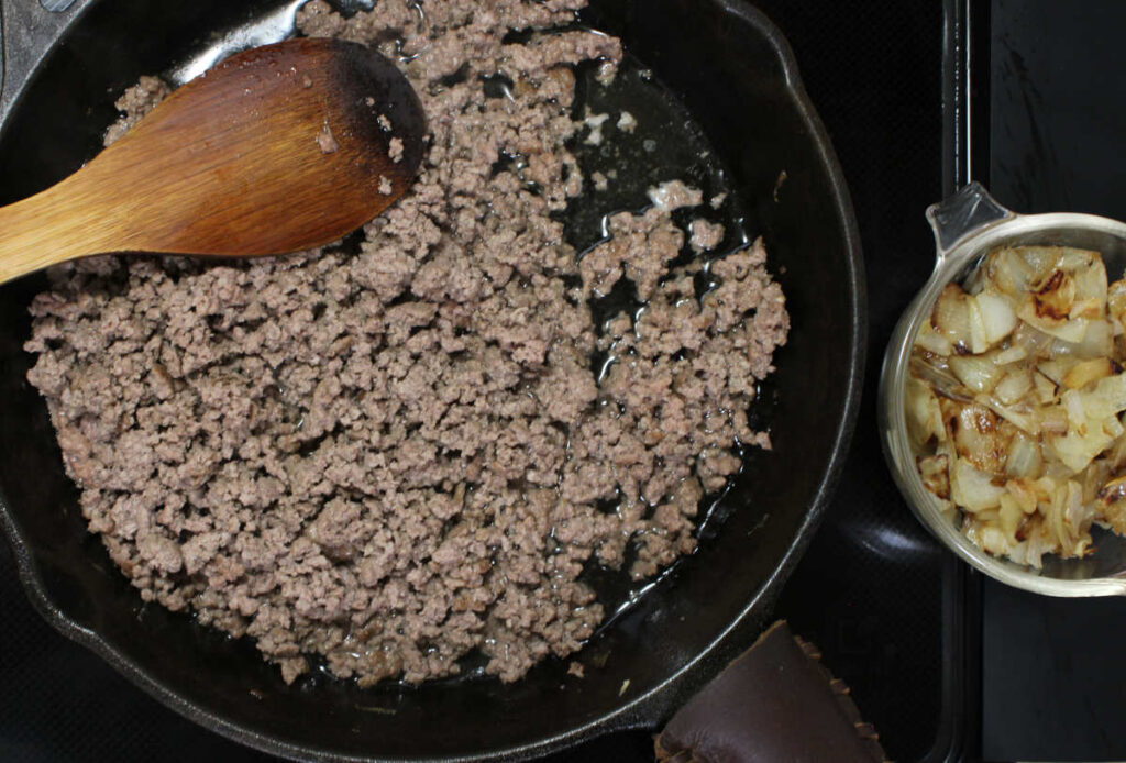 ground beef cooked in a cast iron skillet and a bowl of cooked onions to the side